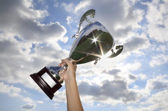 Eine Hand hält einen glänzenden Pokal vor einem wolkigen Himmel.