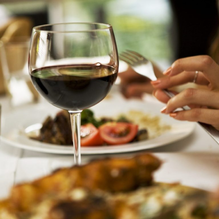 Ein Glas Rotwein mit einer Hand und einem Teller mit Gemüse und Fleisch im Hintergrund.