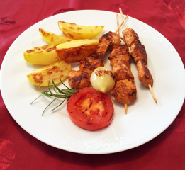 Gegrillte Spieße mit Kartoffelecken, Tomate und Zwiebel auf einem weißen Teller.